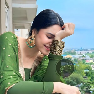 May include: A woman wearing a green and gold patterned dress with green and gold earrings and gold bangles. She is smiling and looking down. The background is a blurry view of a city.