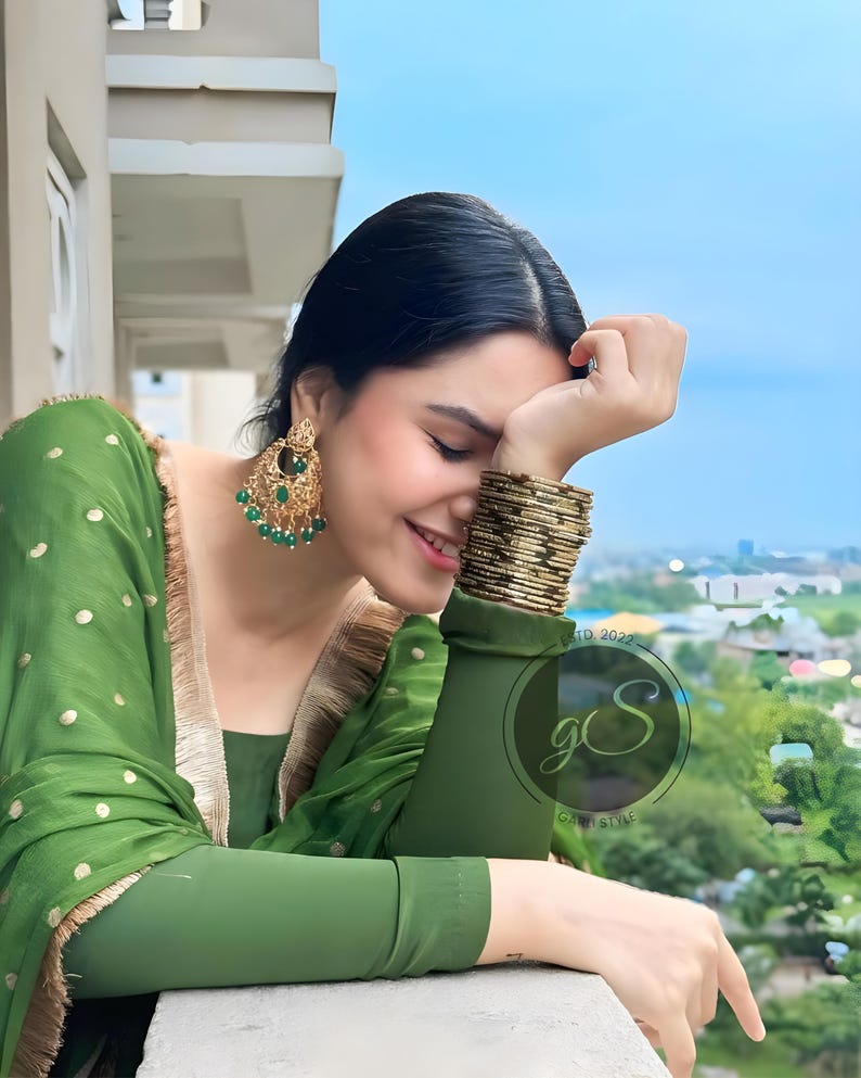 May include: A woman wearing a green and gold patterned dress with green and gold earrings and gold bangles. She is smiling and looking down. The background is a blurry view of a city.