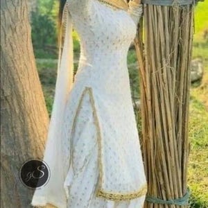 May include: White salwar kameez with gold trim. The outfit features a long-sleeved top and matching pants. The fabric has a subtle pattern. The outfit is paired with a matching scarf. The person is wearing black sandals.