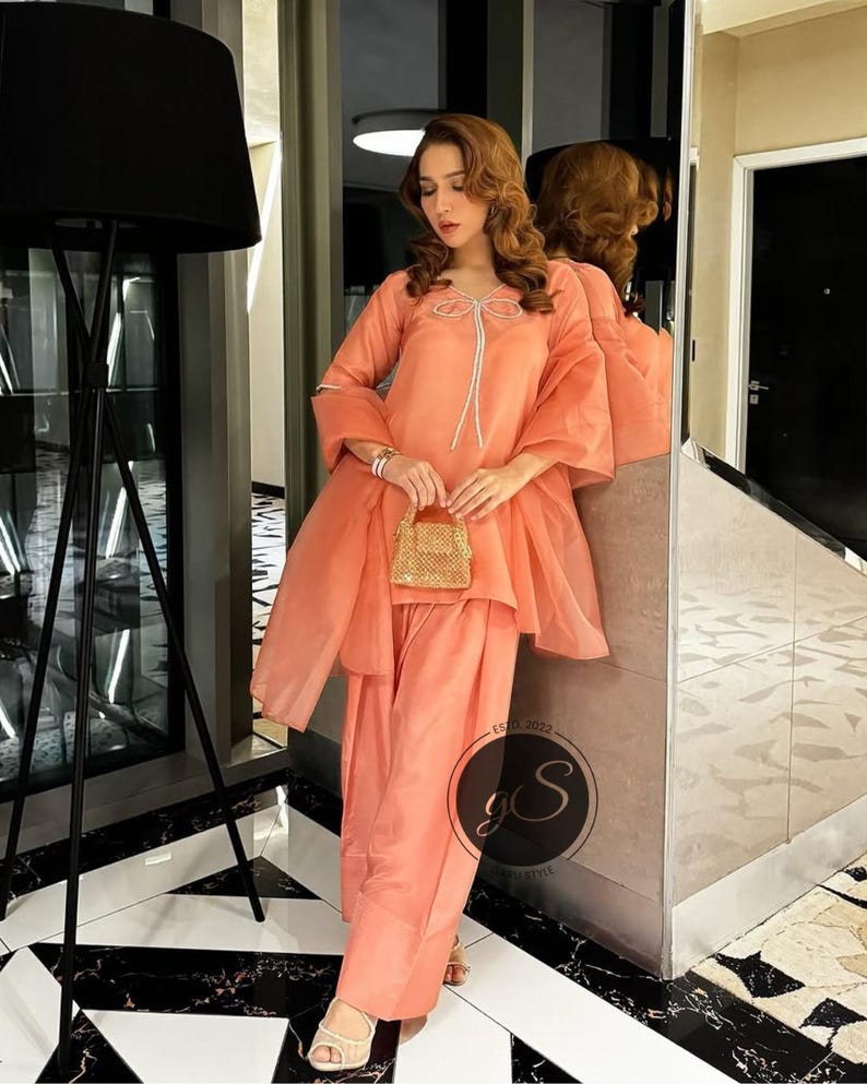Peut inclure: Un salwar kameez de couleur p&ecirc;che avec un d&eacute;tail de n&oelig;ud blanc sur l'encolure. La tenue est associ&eacute;e &agrave; un dupatta assorti et &agrave; un petit sac &agrave; main de couleur or.