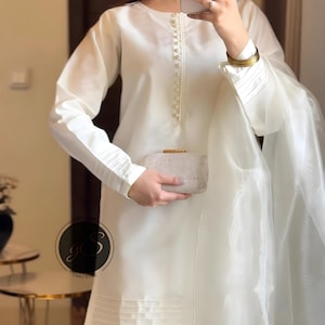 May include: A white long-sleeved tunic with a pearl button closure and a matching white dupatta. The tunic has a pleated hem and the dupatta is sheer. The model is holding a white clutch.