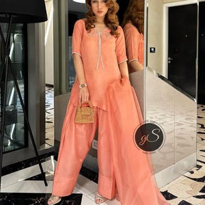 Peut inclure: Une tenue de couleur p&ecirc;che avec un d&eacute;tail de n&oelig;ud sur l'encolure. La tenue comprend un haut &agrave; manches courtes et un pantalon large. Le pantalon a une superposition transparente. Le mannequin tient un petit sac &agrave; main tiss&eacute;.