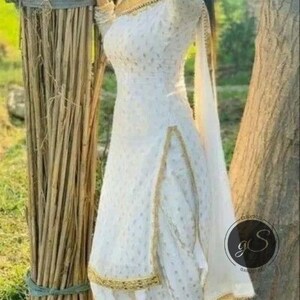 May include: A white salwar kameez with gold trim and a matching dupatta. The outfit features a subtle dotted pattern and is worn with black sandals. The background includes a bundle of sticks and a tree.