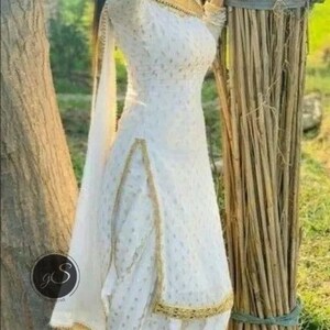 May include: White salwar kameez with gold trim. The outfit features a long-sleeved top and matching pants. The fabric has a subtle pattern. The outfit is paired with a matching scarf. The person is wearing black sandals.