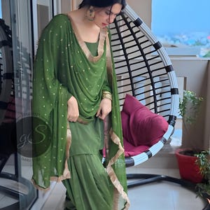 May include: A woman wearing a green salwar kameez with gold embroidery. The outfit includes a long, flowing dupatta with a gold border. The woman is standing in front of a wicker chair.