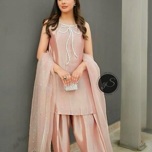 以下が含まれることがあります： 女性がピンクのサルワール・カミーズを着ています。ネックラインにはパールとラインストーンの装飾が施されています。カミーズはボウディテールで、サルワールはゆったりとしたスタイルです。彼女は小さなキラキラしたクラッチを持っています。