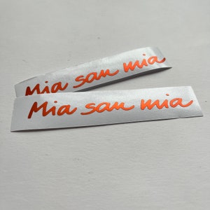 Pode incluir: Duas etiquetas de tecido branco com o texto "Mia san mia" escrito em itálico laranja.