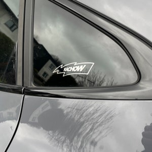 May include: A white vinyl sticker with the word "KACHOW" in a stylized font, with a lightning bolt design, is applied to the rear window of a gray car.