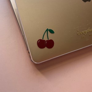 Könnte beinhalten: Ein goldener Laptop mit einem roten Kirsch-Aufkleber auf dem Deckel. Der Aufkleber hat einen grünen Stiel und zwei rote Kirschen.