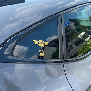 May include: A gold-coloured decal of a winged piston design is affixed to the rear window of a grey car. The decal is visible against the dark-tinted glass, with the car's body reflecting the sky and surrounding environment.