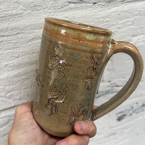 May include: A handmade ceramic mug with a tan and blue-green glaze. The mug features embossed jellyfish designs around the body and a large handle. The mug is held in a hand, showcasing its size and shape.
