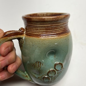 May include: Handmade ceramic mug with a seafoam green glaze and brown accents. The mug features a textured handle and is adorned with seashell designs. The rim and handle are a rich brown color, adding a rustic touch to this unique piece.