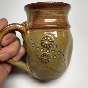 May include: Handmade ceramic mug with a brown rim and handle. The body of the mug is a muted green with embossed floral designs and small blue dots. The mug is held by a hand.