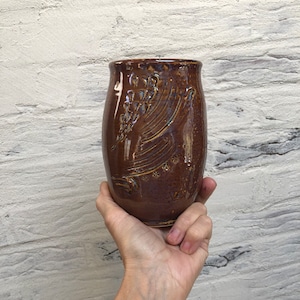May include: A handmade brown ceramic tumbler with a textured surface and carved floral design. The cylindrical cup is held in a hand, showcasing its unique shape and craftsmanship. The background is a white brick wall.
