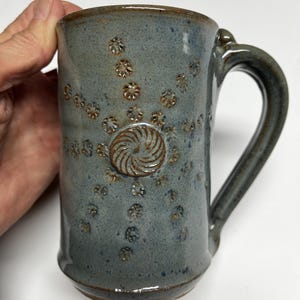 May include: A handmade ceramic mug in a muted blue-gray glaze. The mug features a decorative pattern of small, raised flower-like shapes and a central spiral design. The handle is a matching blue-gray color.