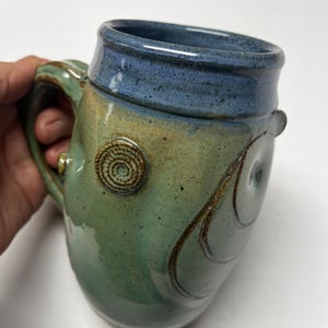 May include: Handmade ceramic mug with a blue rim and handle, and a green and tan body. The mug features spiral and curved line designs. The mug is approximately 4 inches tall and 3 inches wide.