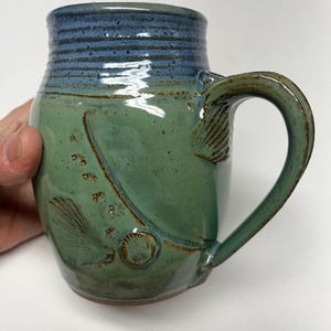 May include: Handmade ceramic mug with a sea-inspired design. The mug features a blue rim and a green body with embossed seashell and bubble details. The handle is curved and the mug has a rustic, textured finish.