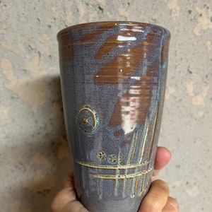 May include: A tall, ceramic mug with a dark purple glaze and gold accents. The mug has a unique design with lines and circles.