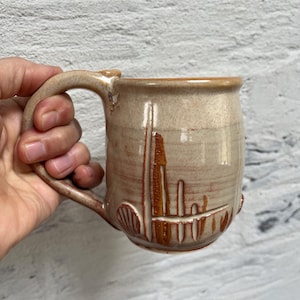 May include: A beige ceramic mug with a brown glaze and a textured design. The mug has a handle and a rounded body.