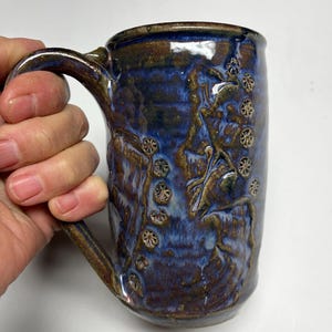 May include: A handmade ceramic mug with a blue and brown glaze. The mug features a textured surface with embossed floral designs and a large handle. The mug is held by a hand, showcasing its size and shape. This unique mug is perfect for coffee or tea.