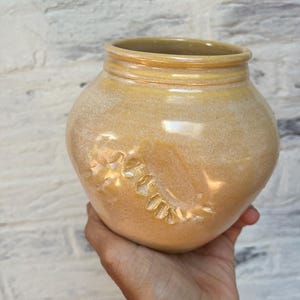 Puede incluir: Un jarrón de cerámica redondo, hecho a mano, con una abertura ancha y un esmalte texturizado de color amarillo-marrón claro. El jarrón presenta un diseño floral tallado en su costado. El jarrón se sostiene en una mano.