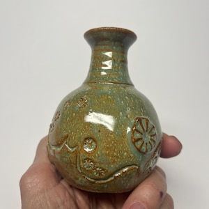May include: A small, handcrafted ceramic vase with a rounded body and a narrow neck. The vase is glazed in shades of green and brown, with embossed floral and geometric patterns. The vase is held in a hand.