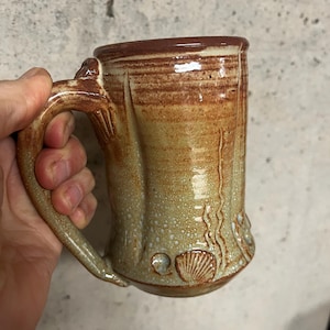 May include: Handmade ceramic mug with a textured, earthy design. The mug features a brown rim and handle, with a gradient of tan and brown tones on the body. Decorative seashell and wave patterns are embossed on the lower portion of the mug.