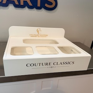 May include: A white rectangular display tray with four recessed compartments and a raised back panel. The back panel features the gold text CULINARY COUTURE, while the front face displays the black text COUTURE CLASSICS with a decorative line element.