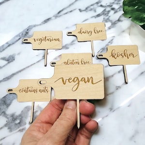 May include: Six wooden signs with different dietary labels: vegetarian, dairy free, gluten free, vegan, kosher, and contains nuts. The signs are on a marble surface.