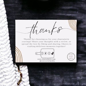 Peut inclure: Une carte de remerciement blanche avec des rayures brunes et dorées. La carte dit "merci" en cursive et comprend le texte "Merci de nous avoir choisis pour vos envies de charcuterie ! Partagez vos pensées avec un avis, ou partagez l’amour en aimant et en partageant. Santé à la création de moments délicieux ensemble !" La carte comprend également des icônes de médias sociaux et le texte "www.yourwebsite.com @yoursocialmedia" et "Votre logo ici".