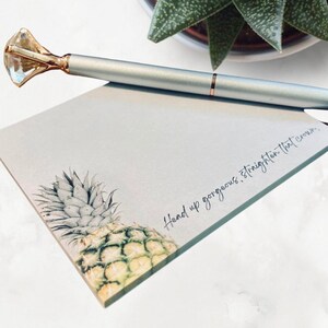 May include: A white notepad with a pineapple illustration and the text "Hand up gorgeous, straighten that crown." A silver pen with a diamond-shaped top is lying on the notepad.