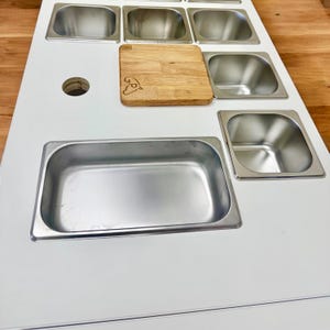 May include: A white serving station with multiple stainless steel containers of varying sizes. A small wooden cutting board with a logo is placed on the surface. A circular hole is visible on the left side.