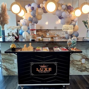 May include: A black catering cart with a Luxe Provisions & Co. logo, topped with food trays and wooden cutting boards. A balloon arch in neutral tones and hanging lights create a festive atmosphere. A water dispenser and glass jars are also visible.