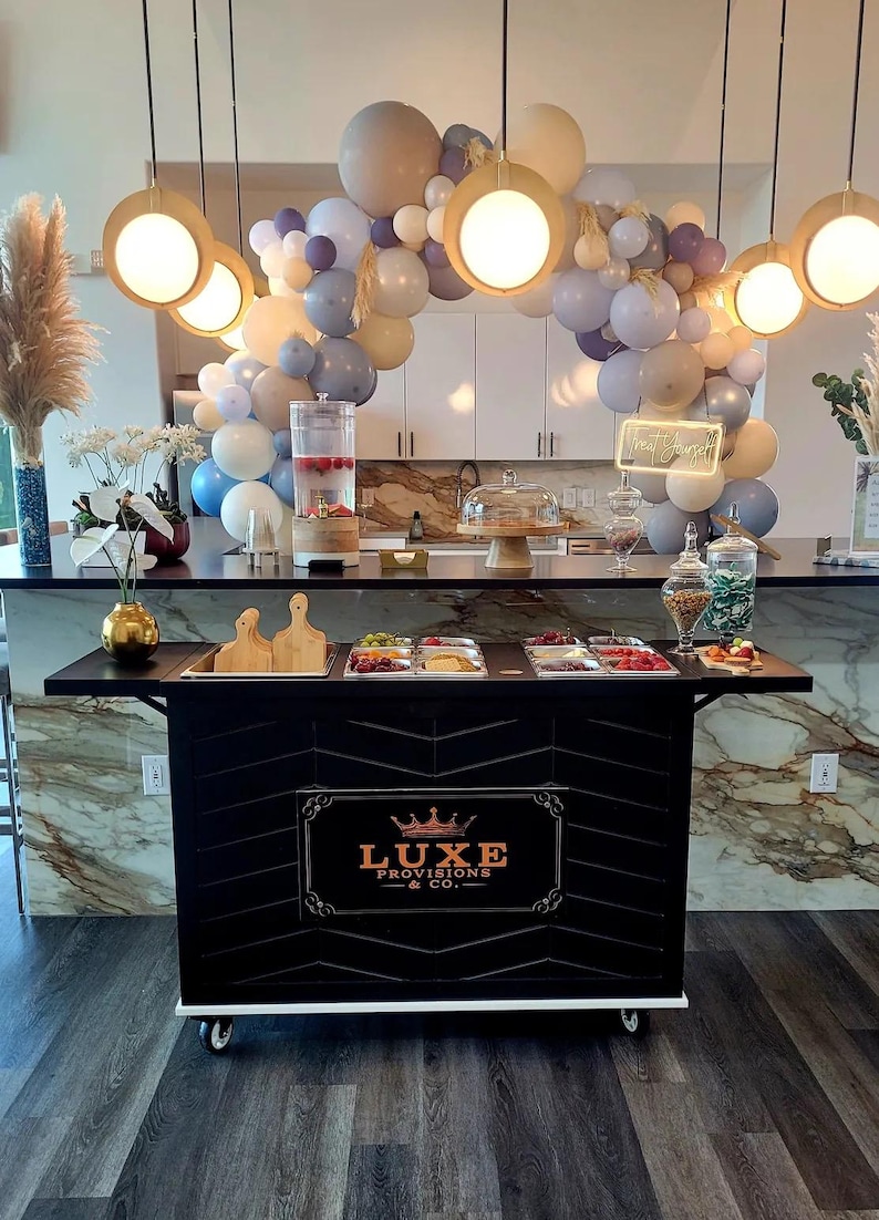 May include: A black catering cart with a Luxe Provisions & Co. logo, topped with food trays and wooden cutting boards. A balloon arch in neutral tones and hanging lights create a festive atmosphere. A water dispenser and glass jars are also visible.