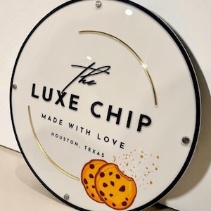 May include: A round white sign with black trim features the text "The Luxe Chip" in black, with "Made with Love" and "Houston, Texas" below. Two chocolate chip cookies are depicted, along with gold accents.