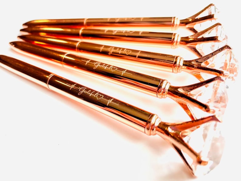 Puede incluir: Cinco bol&iacute;grafos de oro rosa con tapas transparentes en forma de diamante. Cada bol&iacute;grafo est&aacute; grabado con la palabra "Grateful" y un s&iacute;mbolo de coraz&oacute;n.