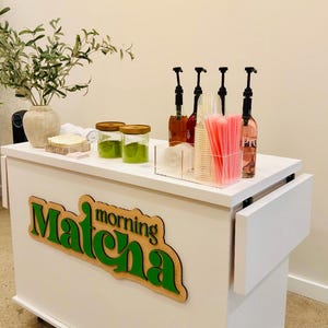 May include: A white mobile cart with the words "Matcha morning" in green and brown. The cart has a countertop with jars of green powder, bottles with pumps, and pink straws. A vase with greenery sits on the left.
