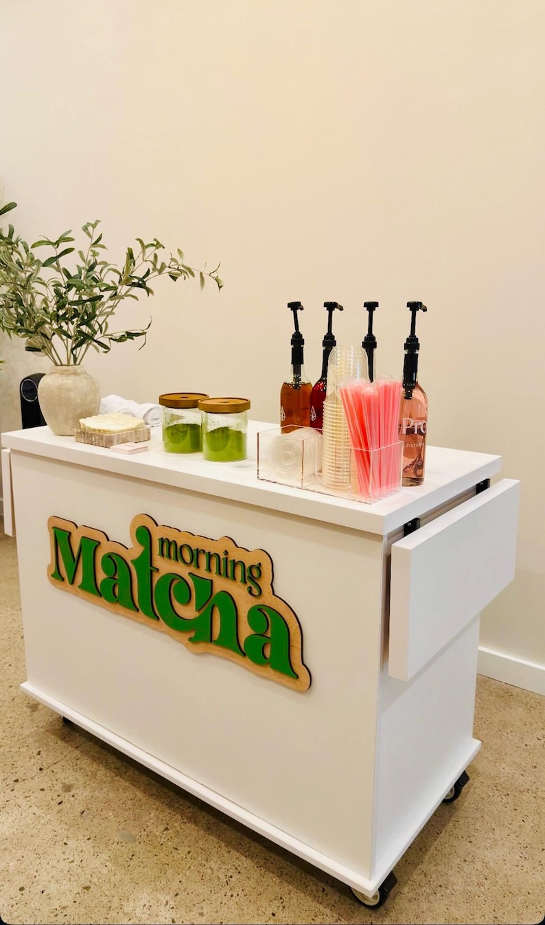 May include: A white mobile cart with the words "Matcha morning" in green and brown. The cart has a countertop with jars of green powder, bottles with pumps, and pink straws. A vase with greenery sits on the left.