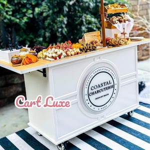 May include: A white charcuterie cart with a wooden countertop, displaying various food items. The cart features the text "Coastal Charcuterie Newport Beach & More" in a circular design. The cart is on wheels and sits on a black and white striped rug.