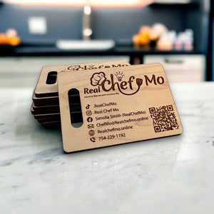 May include: A wooden business card with the text "Real Chef Mo" and a chef hat logo. The card includes contact information, social media handles, and a QR code.