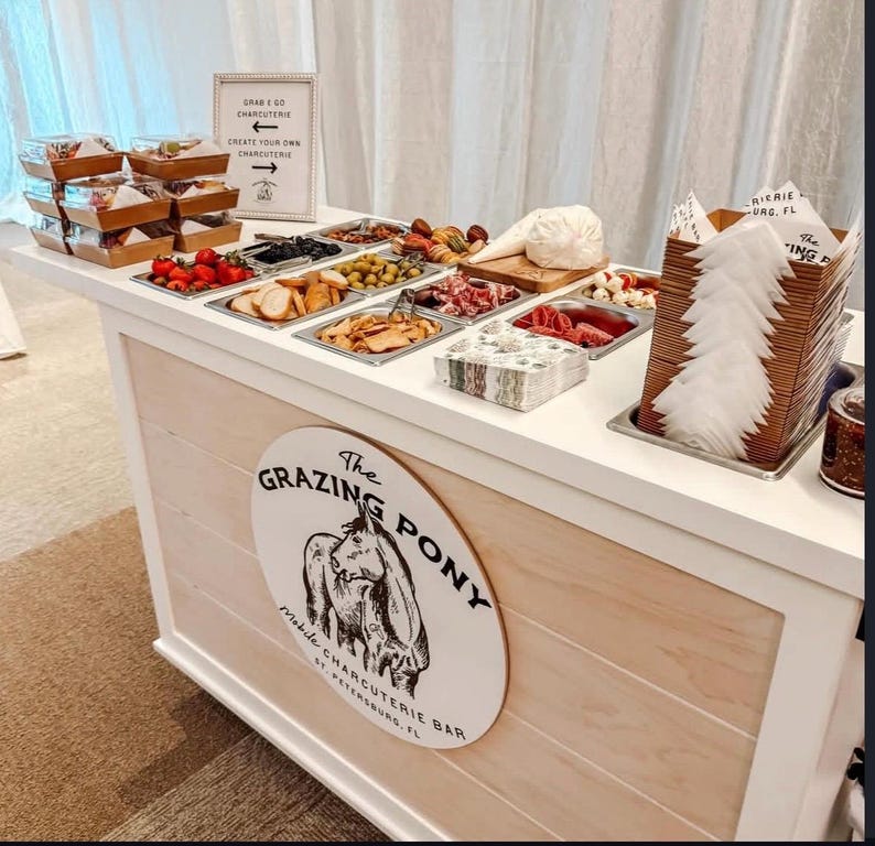 May include: A charcuterie bar with various food items in metal trays, including olives, meats, and cheeses. The bar features a sign that reads "The Grazing Pony" and "Mobile Charcuterie Bar, St. Petersburg, FL."