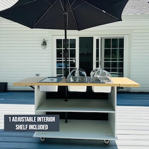May include: A white and tan outdoor kitchen cart with a black umbrella. The cart has a light wood-colored countertop, two clear plastic food covers, and a built-in sink. The text "1 ADJUSTABLE INTERIOR SHELF INCLUDED" is visible.