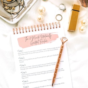 Puede incluir: Un diario de gratitud rosa y blanco con una encuadernaci&oacute;n en espiral dorada y un bol&iacute;grafo de color rosa dorado con una parte superior en forma de diamante. El diario tiene el texto "The 2 Minute Intentionally Grateful Routine" escrito en la portada.