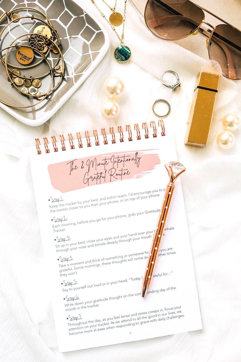 Puede incluir: Un diario de gratitud rosa y blanco con una encuadernaci&oacute;n en espiral dorada y un bol&iacute;grafo de color rosa dorado con una parte superior en forma de diamante. El diario tiene el texto "The 2 Minute Intentionally Grateful Routine" escrito en la portada.