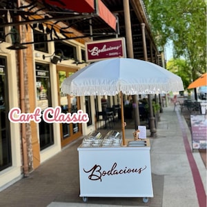 May include: A white cart with the text "Bodacious" in a cursive font, topped with a white umbrella with fringe. The cart has two clear containers and a wooden counter. The text "Cart Classic" is in pink cursive.