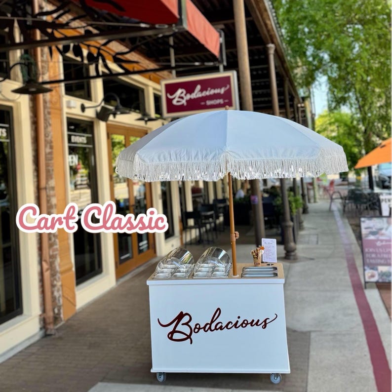 May include: A white cart with the text "Bodacious" in a cursive font, topped with a white umbrella with fringe. The cart has two clear containers and a wooden counter. The text "Cart Classic" is in pink cursive.