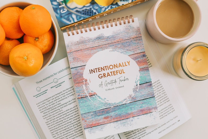 Journal Intentionally Grateful, a gratitude practice journal, laying on top of another book near oranges and coffee