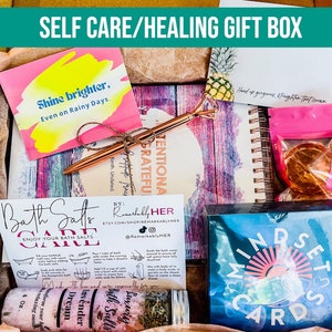 Puede incluir: Una caja de regalo de autocuidado llena de art&iacute;culos como sales de ba&ntilde;o, un diario, tarjetas de afirmaci&oacute;n y una tarjeta de cita motivacional. La caja est&aacute; decorada con una tarjeta rosa y amarilla que dice "Shine brighter, Even on Rainy Days."