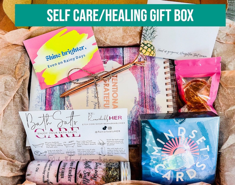 Puede incluir: Una caja de regalo de autocuidado llena de art&iacute;culos como sales de ba&ntilde;o, un diario, tarjetas de afirmaci&oacute;n y una tarjeta de cita motivacional. La caja est&aacute; decorada con una tarjeta rosa y amarilla que dice "Shine brighter, Even on Rainy Days."