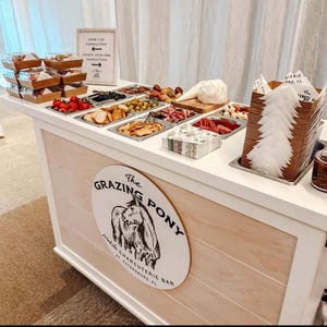 May include: A charcuterie bar with various food items in stainless steel trays. The bar features a sign that says "The Grazing Pony" and "Mobile Charcuterie Bar". There are also pre-packaged charcuterie boxes and a sign that says "Grab & Go Charcuterie".